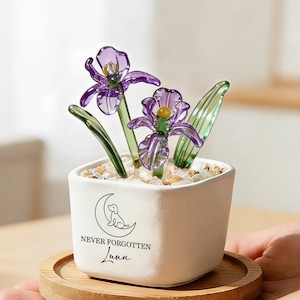 Puede incluir: Una pequeña maceta de cerámica blanca cuadrada que contiene dos flores de iris de vidrio morado con tallos verdes y guijarros claros. La maceta presenta un dibujo lineal negro de un perro sobre una luna creciente con el texto NEVER FORGOTTEN Luna.