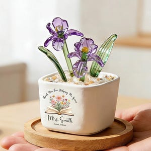 May include: A small white square ceramic planter containing two purple glass flowers with green stems and clear pebbles. The planter features a printed graphic of an open book with colourful flowers and the text "Thank You For Helping Me Grow", "Mrs. Smith", and "Love, Oliver". It sits on a round wooden coaster.