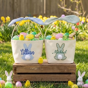 May include: Two cream Easter baskets with gingham bunny ear handles, filled with eggs, chicks, and a bunny figurine. One basket has "Lillian" and a blue bunny, the other has "Michael" and a green bunny. Set on a wooden crate in a garden.