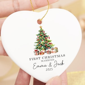 Könnte beinhalten: Weißer herzförmiger Weihnachtsschmuck mit goldenem Band. Der Schmuck zeigt eine Aquarellillustration eines geschmückten Weihnachtsbaums und Geschenken. Der Text lautet "First Christmas Married Emma & Jack 2025".