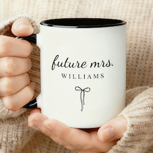 May include: White ceramic mug with a black interior and handle. The mug features the text "future mrs. WILLIAMS" in a stylish script, along with a small bow illustration. The mug is held by a person wearing a beige jumper.