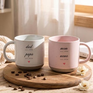 May include: Two ceramic mugs with speckled finishes, one white and one pink. The white mug features the text dad est.1994, papa est.2026, and a small red heart. The pink mug features the text mom est.1996, nana est.2026, and a small red heart.