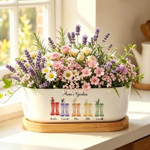 May include: A white oval ceramic planter on a wooden base filled with lavender, daisies, and pink flowers. The planter features the text "Mom's Garden" above a graphic of five pairs of colourful rain boots with names underneath each pair.