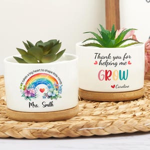 Peut inclure: Deux pots de plantes en céramique blanche avec des bases en bambou, chacun contenant une plante succulente. L'un des pots présente un motif arc-en-ciel avec le texte "It takes a big heart to shape little minds" et "Mrs. Smith". L'autre dit "Thank you for helping me GROW" et "Caroline."