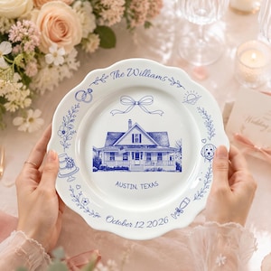 May include: A white ceramic plate with a scalloped edge featuring a blue ink illustration of a house in the centre. Text reads The Williams, Austin, Texas, and October 12 2026. Decorative blue icons include a bow, rings, a cake, and a dog.