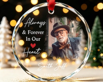 May include: A circular glass ornament with beveled edges and a red ribbon hanger. It features a photograph of an older person wearing a hat and glasses, the text "Always & Forever In Our Heart", the name "Smith Jeff", and the dates "1941-2026".