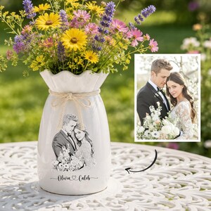 May include: A white ceramic vase with a ruffled rim and a jute twine bow, featuring a black and white line art illustration of a couple and the text "Olivia & Caleb" printed on the front, filled with a bouquet of yellow, pink, and purple wildflowers.