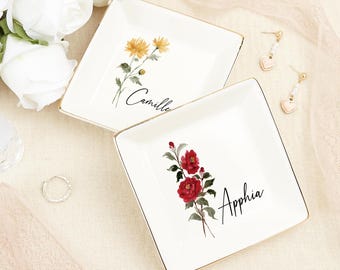 Könnte beinhalten: Zwei weiße Keramik-Schmuckschalen mit goldener Verzierung. Eine Schale zeigt eine Aquarell-Illustration von gelben Blumen mit dem Namen "Camille" darunter. Die andere Schale zeigt eine Aquarell-Illustration von roten Blumen mit dem Namen "Apphia" darunter.