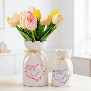 May include: Two white ceramic vases with scalloped rims and cream ribbon ties. One vase holds a bouquet of pink and yellow tulips. The vases have heart designs with the words "Gran" and "Mama" and names.