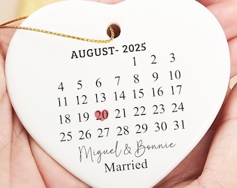 May include: White heart-shaped ceramic ornament with a calendar for August 2025, with the 20th highlighted in red. The text "Miguel & Bonnie Married" is written below the calendar.