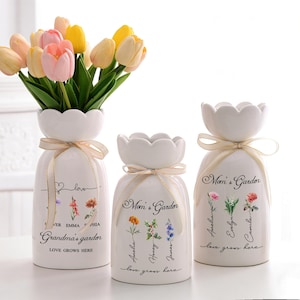 May include: Three white ceramic vases with floral designs and the text "Mom's Garden" and "Grandma's Garden". Beige ribbons adorn the vases, which hold colourful tulips. The text "Love Grows Here" is also visible.