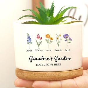 Op de afbeelding: Een witte keramische plantenpot met een bamboe basis. De pot heeft vijf bloemen met namen onder elke bloem: Adele, Winnie, Alani, Bonnie en Jacob. De tekst "Grandma's Garden Love Grows Here" is op de pot gedrukt.