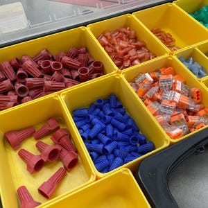 May include: A yellow organizer filled with various electrical wire connectors. The compartments hold red, blue, orange, and green connectors. The red connectors are cone-shaped, while the blue ones are cylindrical. The orange connectors are rectangular.