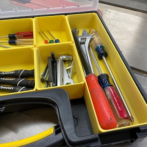 Op de afbeelding: Een zwarte en gele gereedschapskist gevuld met diverse handgereedschappen. De gereedschappen omvatten schroevendraaiers, sleutels en inbussleutels. De schroevendraaiers hebben rode, blauwe en zwarte handvatten. De gereedschapskist is open en toont de georganiseerde compartimenten.