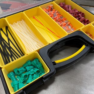 May include: A black and yellow organizer filled with various electrical components. The compartments hold black and white cable ties, orange and red wire connectors, and green wire nuts. A yellow handle is visible.