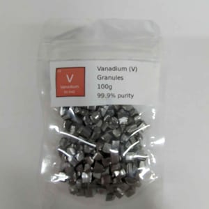 Può includere: Un sacchetto di plastica trasparente contenente granuli di vanadio grigio scuro. Un'etichetta indica che il contenuto è di 100 g di vanadio (V) puro al 99,9%. L'etichetta include anche il simbolo dell'elemento e il numero atomico.