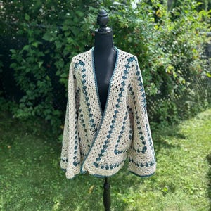 May include: A beige crochet cardigan with dark teal trim and accents. The open-front sweater is displayed on a black dress form against a backdrop of green foliage. The crochet pattern features a repeating geometric design.