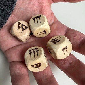 May include: Four wooden dice with black etched symbols are held in a hand. The dice are light brown and have various geometric designs on each face. The dice appear to be new and unused.