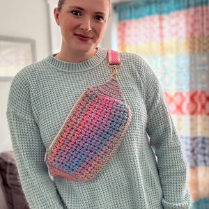 May include: A woman wearing a light blue sweater and blue jeans is wearing a rainbow crocheted fanny pack with a pink and white strap.