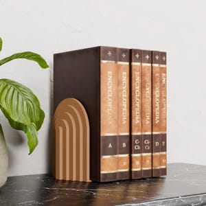 May include: A set of brown leather-bound encyclopedias with gold lettering and a decorative arch-shaped bookend. The books are labeled "ENCYCLOPEDIA" and have volume letters and numbers.