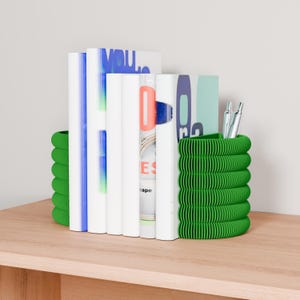 Puede incluir: Un organizador de escritorio verde, estilo acordeón, sostiene libros y bolígrafos sobre una superficie de madera. El organizador tiene un diseño acanalado y está colocado a ambos lados de los libros. Los libros tienen cubiertas coloridas y los bolígrafos son plateados.