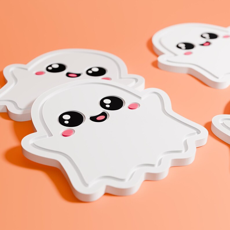 May include: White ghost-shaped coasters with black eyes, a smiling mouth, and pink cheeks. The coasters are arranged on an orange surface. These decorative coasters are perfect for Halloween or spooky-themed decor.