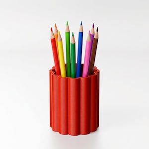 May include: A red pencil holder with a wavy top holds a collection of colorful pencils. The pencils are in various colors, including red, yellow, green, blue, pink, and brown. The pencils are arranged upright in the holder, creating a vibrant display.