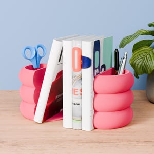 Puede incluir: Organizadores de escritorio rosas con un diseño apilado y redondeado. Uno contiene tijeras azules, el otro bolígrafos. Libros blancos con detalles coloridos se interponen entre los organizadores. Una planta está en el fondo.
