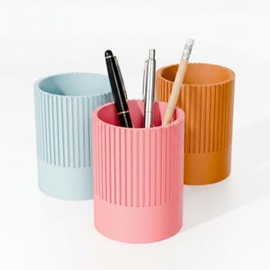 May include: Three colorful pen holders in pastel shades of blue, pink, and orange. Each cylindrical holder has vertical ribbed detailing and a solid base. Pens and a pencil are inside the pink holder.
