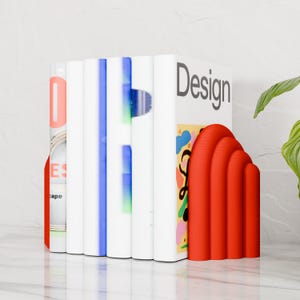 Op de afbeelding: Een stapel boeken met een rode, gebogen boekensteun. De boeken hebben witte ruggen met blauwe en groene accenten. De boekensteun is felrood en heeft een gebogen ontwerp. Het woord "Design" is zichtbaar.