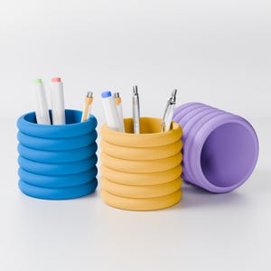 Op de afbeelding: Drie kleurrijke pennenbakjes in blauw, geel en paars. De blauwe en gele houders staan rechtop en zijn gevuld met pennen en stiften. De paarse houder ligt op zijn kant. De houders hebben een uniek, gestapeld ringontwerp.