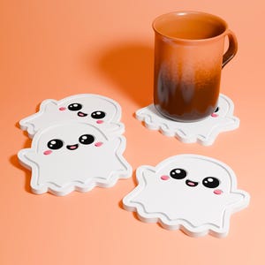 May include: Four white ghost-shaped coasters with black eyes, pink cheeks, and a smiling mouth. A brown mug rests on one coaster. The coasters are on an orange background.