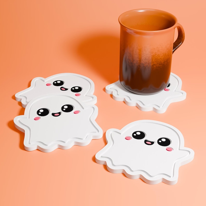 May include: Four white ghost-shaped coasters with black eyes, pink cheeks, and a smiling mouth. A brown mug rests on one coaster. The coasters are on an orange background.