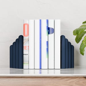 May include: A pair of navy blue bookends with a scalloped design, holding a row of white books. The book on the left has the word "scape" visible. The books are on a white surface.