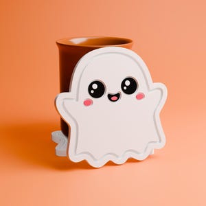 May include: A white, ghost-shaped coaster with a cute face, featuring large black eyes, pink cheeks, and a smiling mouth. The coaster is in front of a brown mug, set against an orange background. A fun Halloween or spooky season themed item.