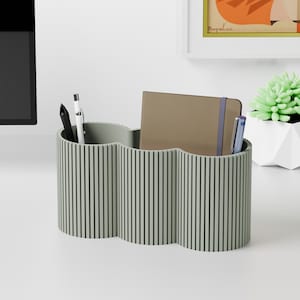 Puede incluir: Un organizador de escritorio verde salvia con un diseño ondulado y líneas verticales. El organizador contiene un cuaderno marrón, bolígrafos y un bolígrafo. El organizador está sobre una superficie blanca.