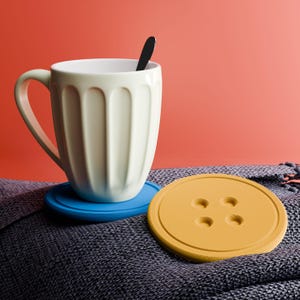 Puede incluir: Una taza crema con una cuchara negra dentro descansa sobre un posavasos azul. Un posavasos amarillo en forma de botón está al lado. Los artículos están sobre una superficie texturizada gris oscuro contra un fondo coral.
