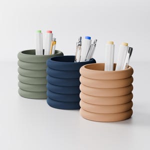 May include: Three cylindrical pen holders in olive green, navy blue, and tan. Each holder is made of stacked, rounded rings. White pens and pencils with colored accents are inside the holders. The holders are on a white surface.