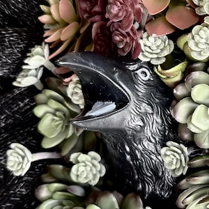 May include: A black raven sculpture with an open beak, surrounded by a variety of succulents in shades of green, pink, and burgundy. The raven's textured feathers and open mouth create a striking contrast with the soft, fleshy plants.