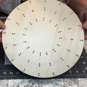 May include: A round wooden board with holes drilled in a circular pattern. The board is light brown and has a smooth surface.