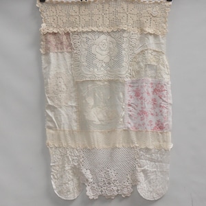 Peut inclure: Textile patchwork crème, probablement un rideau ou une tenture murale, composé de divers tissus. Il présente de la dentelle, du crochet et des motifs floraux, avec un bord inférieur festonné. Suspendu à un cintre noir.
