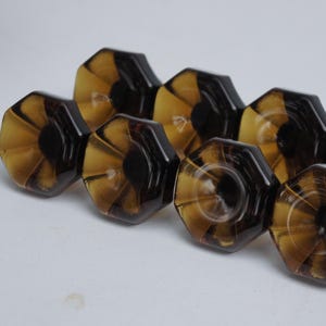 Puede incluir: Ocho tiradores hexagonales para armarios en marrón oscuro y ámbar. Cada tirador presenta un diseño radial que emana de un círculo central marrón oscuro. Los tiradores están dispuestos en fila.