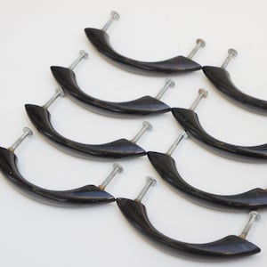 Op de afbeelding: Een verzameling van acht zwarte, gebogen kastgrepen met zilveren schroeven. De handgrepen hebben een glanzende afwerking en zijn gerangschikt op een wit oppervlak. Deze vintage handgrepen zijn geschikt voor meubelrestauratieprojecten.