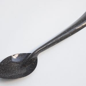 May include: A black and white speckled spoon with a long handle and a rounded bowl. The spoon has a small hole at the end of the handle. The spoon is made of metal and is likely used for cooking or serving food.