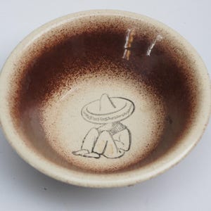 May include: A round ceramic bowl with a cream-colored base and a dark brown speckled gradient finish along the interior rim. At the center of the bowl is a black line drawing of a figure sitting down while wearing a large, wide-brimmed sombrero hat.