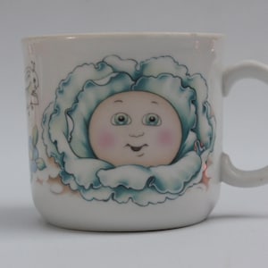 Könnte beinhalten: Weiße Keramik-Tasse mit einer skurrilen Illustration eines Babygesichts, umgeben von einem blau-grünen Kohl. Die Tasse hat einen gebogenen Henkel und ist für Heißgetränke geeignet. Das Design erinnert an ein Vintage-Sammlerstück.