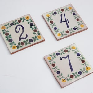 May include: Three square ceramic tiles with hand-painted numbers 2, 4, and 7. Each tile has a cream-colored background, a dark blue number, and is framed by a floral border with green leaves, yellow, purple, and red flowers.