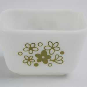 May include: A square, white glass dish with a floral design in olive green. The flowers and dots are arranged on the side of the dish. The dish is likely vintage and suitable for serving or storage.