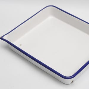 May include: A white rectangular enamel tray with a blue trim. The tray has a slightly raised edge and a smooth, glossy surface. The corners are rounded. The tray is empty and clean.