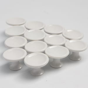 May include: A collection of white ceramic cabinet knobs. Each knob features a round, slightly concave top and a short, pedestal-style base. The knobs are arranged on a white surface.